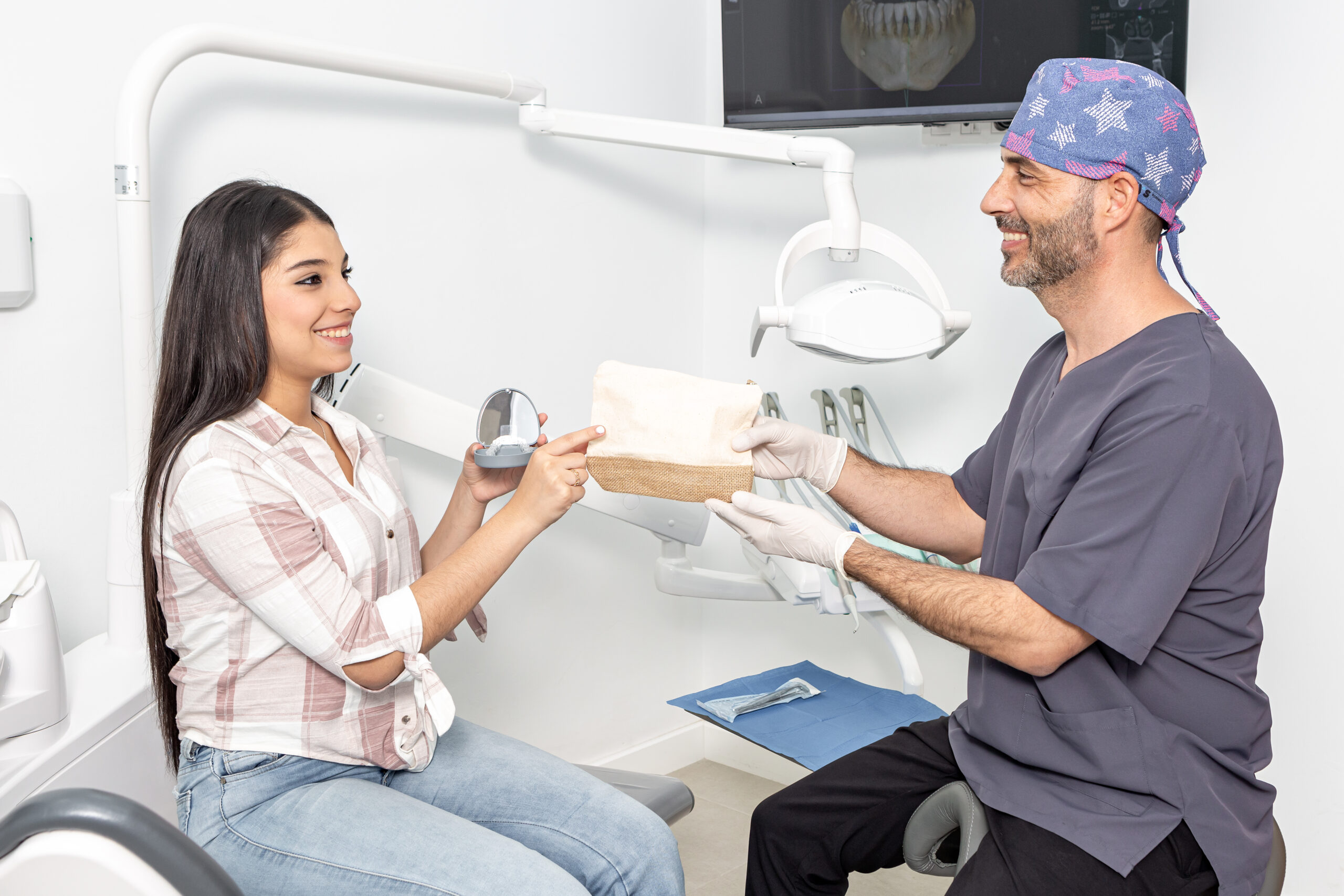 Side view of delighted ethnic teen girl with long dark hair in casual clothes with aligner box in hand taking prescription from hands of smiling male dentist during check up in clinic